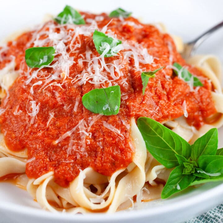 Roasted tomato pasta sauce over tagliatelle in white bowl with fork garnished with Parmesan cheese and fresh basil.