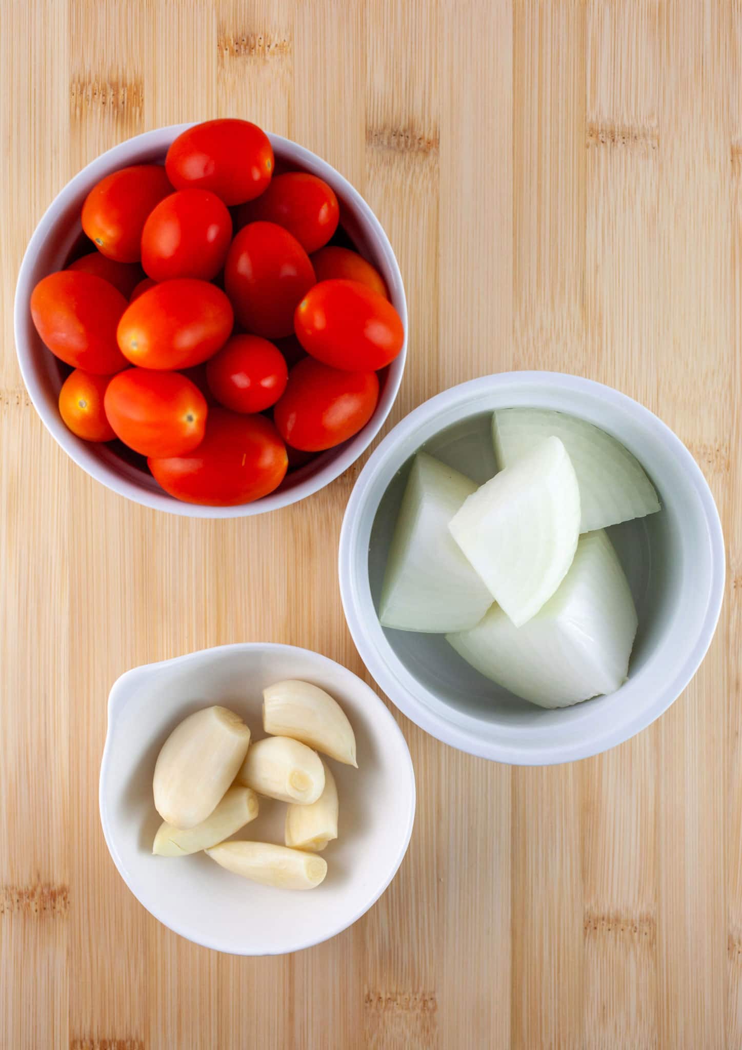 Garlic cloves, chopped onions, and grape tomatoes in small white bowls on wooden cutting board.