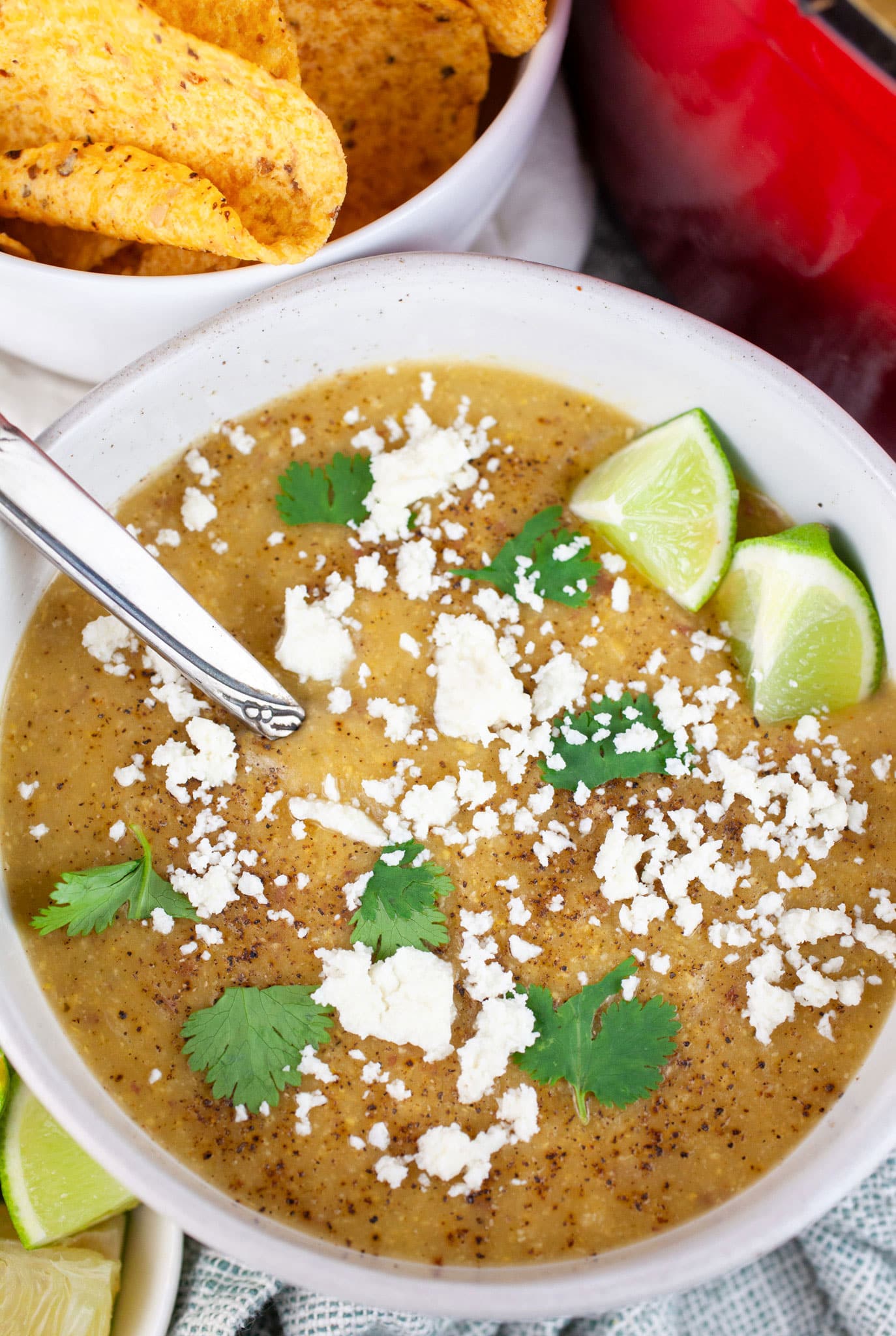 Southwest corn potato chowder garnished with Queso fresco, cilantro, and lime wedges in white bowl with spoon next to bowl of tortilla chips.