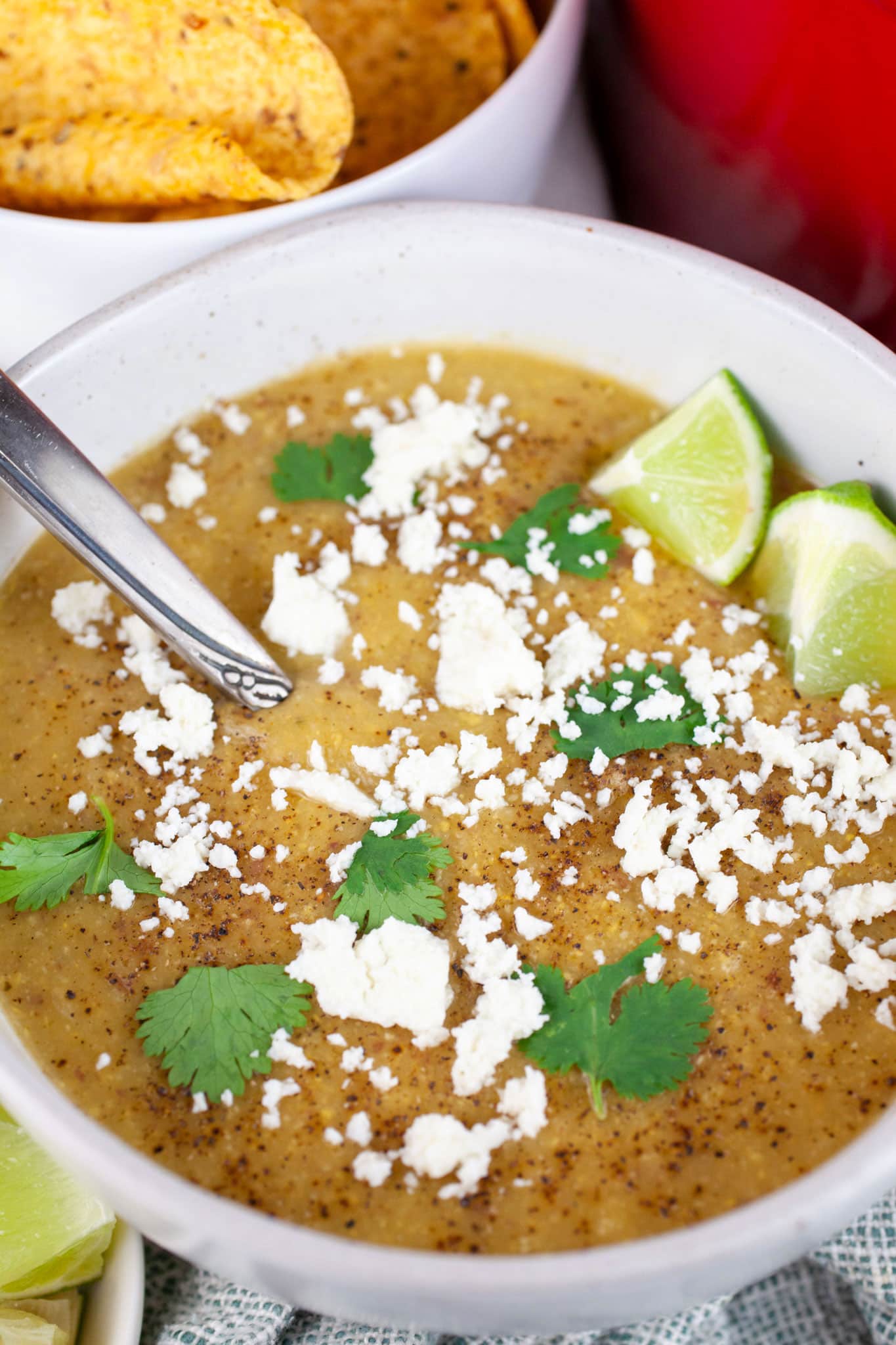Southwest potato corn chowder in white bowl with spoon garnished with Queso fresco, lime wedges, and cilantro next to bowl of tortilla chips.