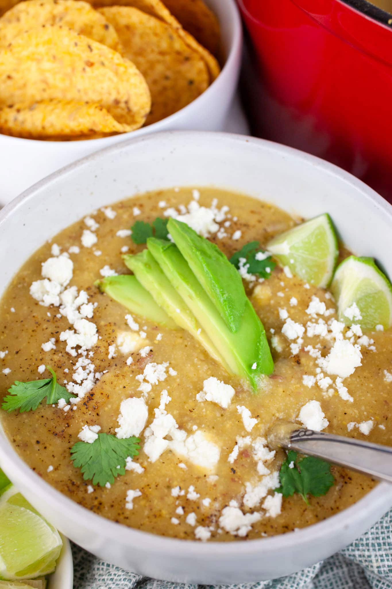 Southwest potato corn chowder in white bowl with spoon garnished with avocadoes, Queso fresco, cilantro, and lime wedges next to bowl of tortilla chips.