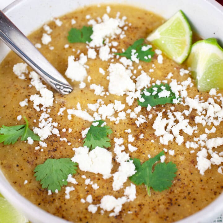 Southwest potato corn chowder in white bowl with spoon garnished with Queso fresco, cilantro, and lime wedges.