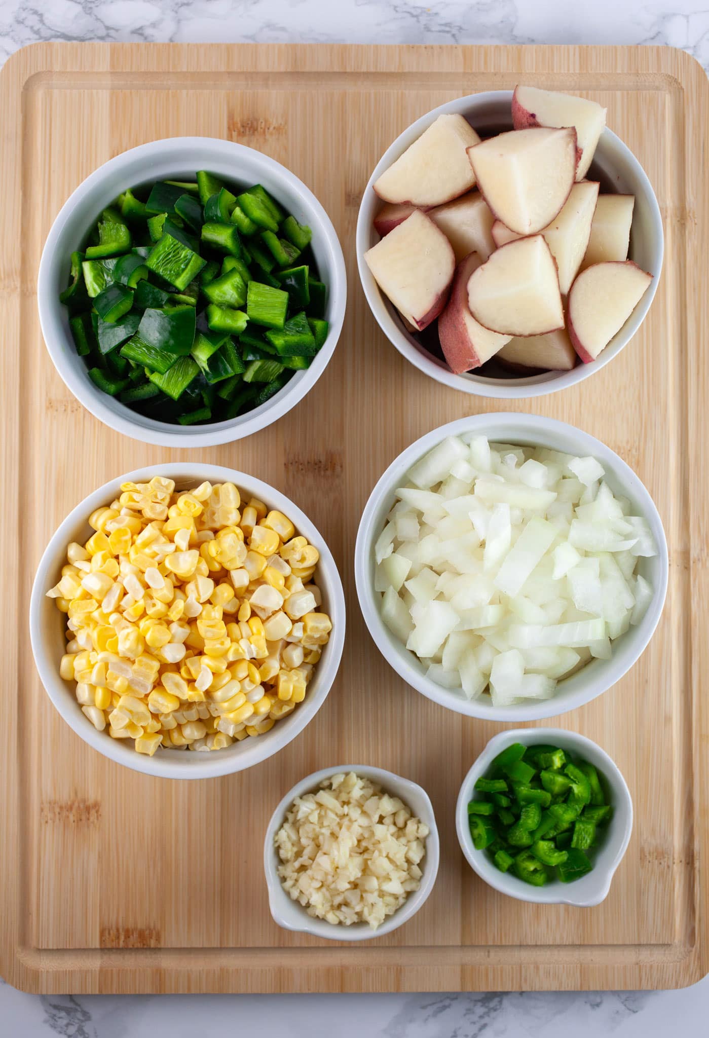 Minced garlic, onions, and jalapeno and poblano peppers and sweet corn kernels and diced potatoes in small white bowls on wooden cutting board.