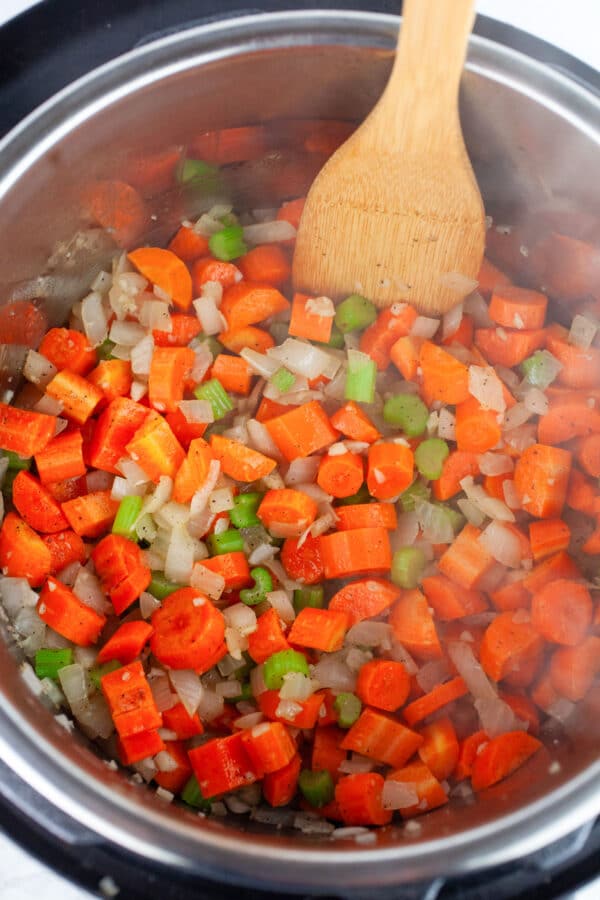 Minced garlic, onions, and celery and sliced carrots sauteed in Instant Pot with wooden spatula.