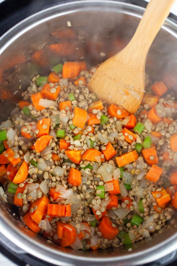Minced garlic, onions, and celery, sliced carrots, and dried lentils sauteed in Instant Pot with wooden spatula.