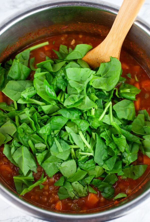 Chopped spinach added to lentil soup in Instant Pot with wooden spatula.