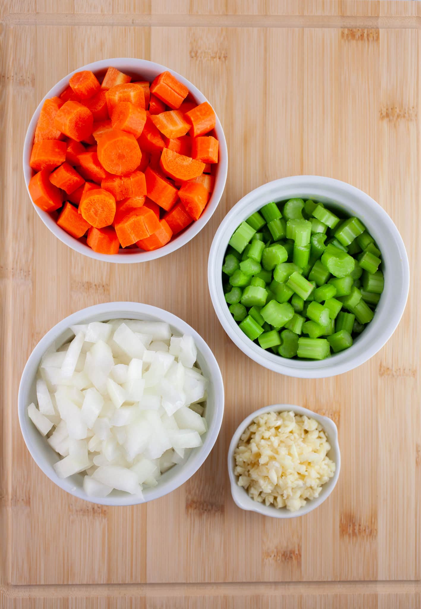 Minced garlic, onions, and celery and chopped carrots in small white bowls on wooden cutting board.